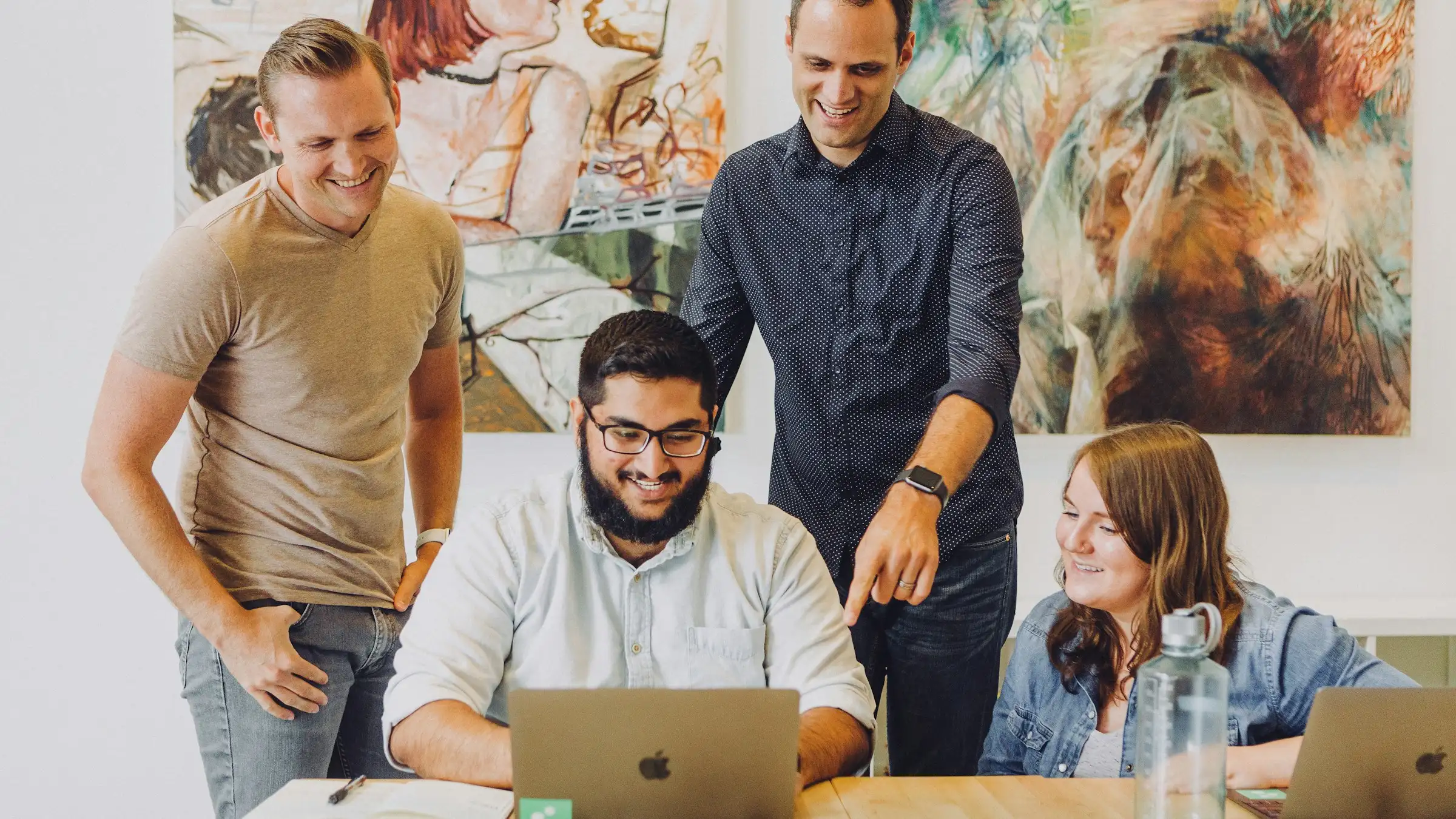 Four coworkers gathered around a laptop at a wooden table, reviewing work on screen.