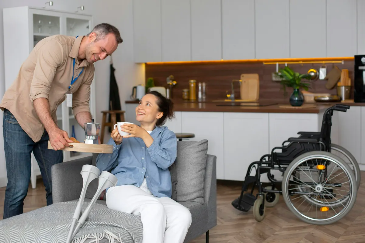 A support worker hands a cup of coffee to a woman seated in her living room, her wheelchair beside the sofa.