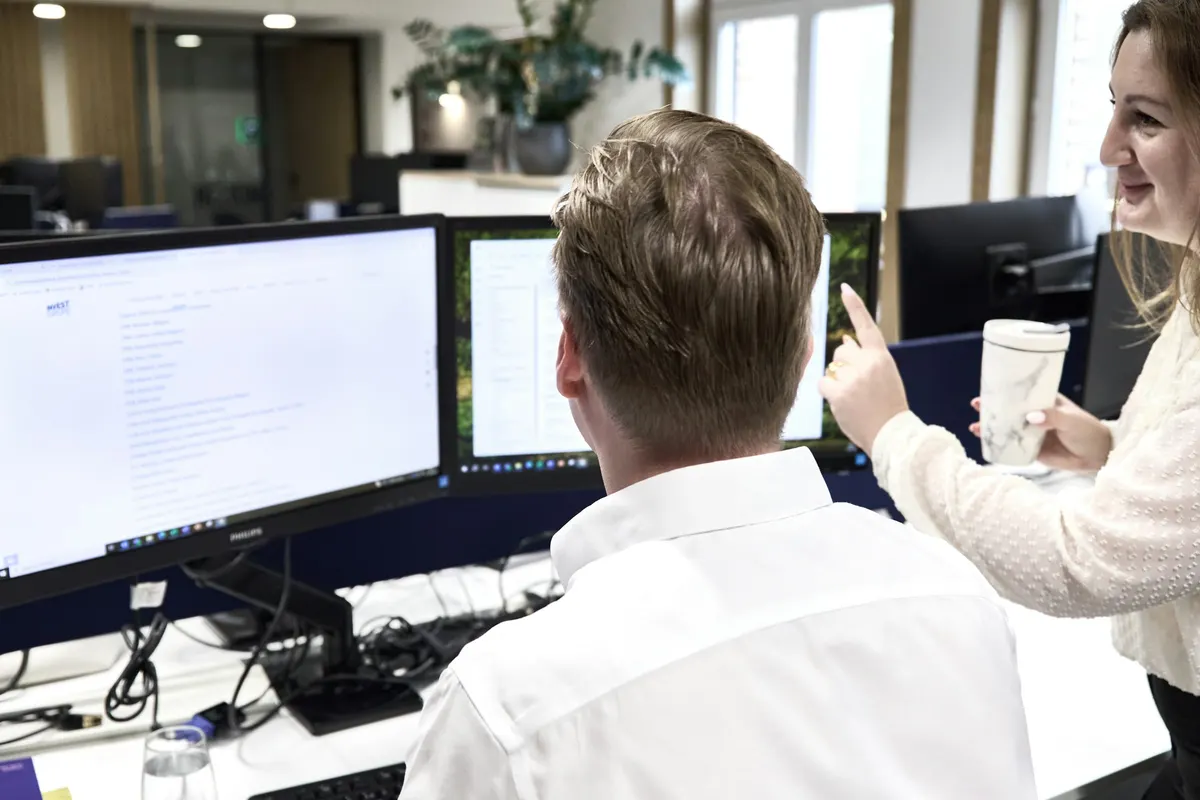 Two colleagues at a desk with several monitors, one pointing at the screen.