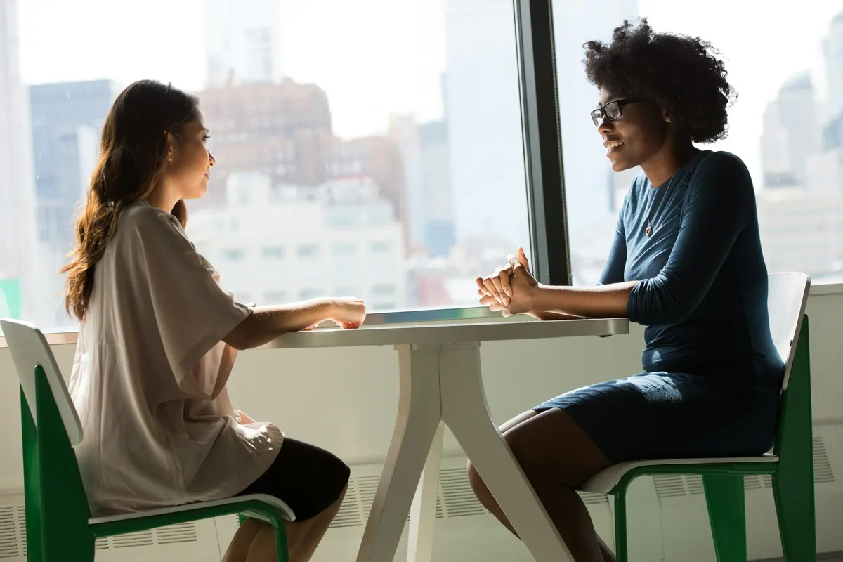 Two women in conversation across a small table by a window.